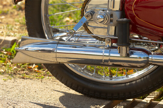 Close-up of rear motorcycle wheel showing chrome exhaust, sprocket, chain, and tire on sunlit pavement with reflected surroundings.