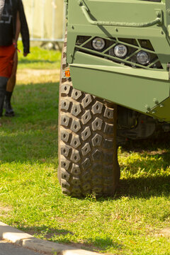 Close-up of front wheel and headlight assembly of green armored vehicle parked on grass with partial pedestrian in background.