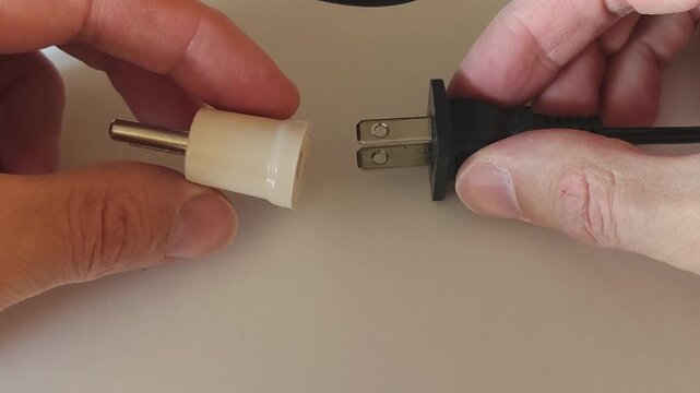 Close-up of hands holding an electrical plug and socket. Connecting a black power plug to a beige adapter, top view, white background.