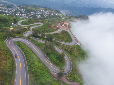 Drone shot of S-curve winding asphalt road along steep mountain ridge surrounded by lush green nature and sea of fog. Road trip, travel destination, and transportation concept.
