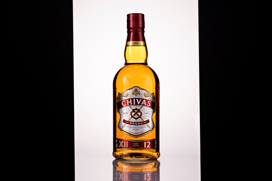 Front view of a Chivas Regal 12 Year Old blended Scotch whisky bottle, backlit to reveal its golden amber colour, with burgundy and gold labels and a reflection on a glossy surface against a white bac