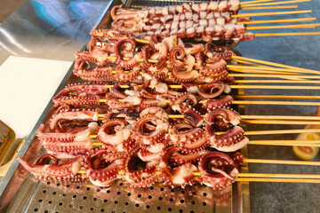 Rows of raw octopus tentacles skewered on wooden sticks rest atop perforated metal tray at Asian street food market stall ready for grilling, offering fresh seafood delicacies to hungry visitors © RollAndSnap