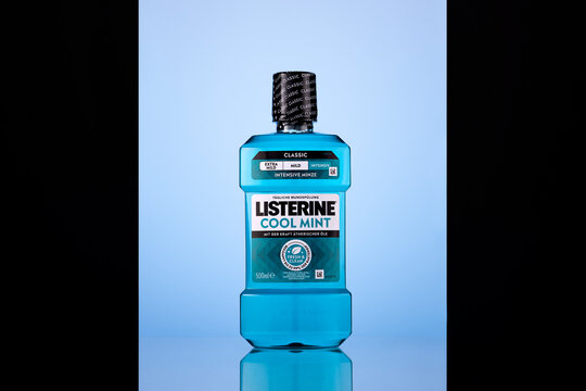 Front view of a Listerine Cool Mint Classic 500 ml mouthwash bottle with turquoise liquid, a black cap, and German-language labels. Backlit studio composition on a reflective surface against a pale bl