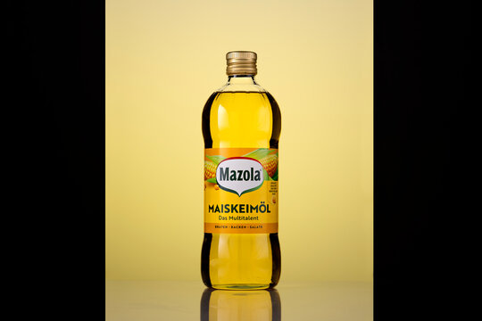 Front view of a Mazola corn oil bottle with a gold-coloured screw cap and German Maiskeim&ouml;l label. Backlighting illuminates the golden cooking oil against a yellow background framed by black panels, w