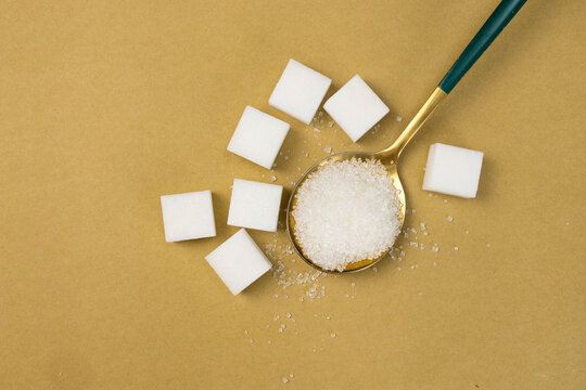 A gold spoon filled with white granulated sugar surrounded by sugar cubes on a beige background