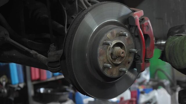 Auto mechanic cleans rust off a car brake caliper bracket with a rotary wire tool. Compressed air blow gun removes dust during workshop service maintenance.