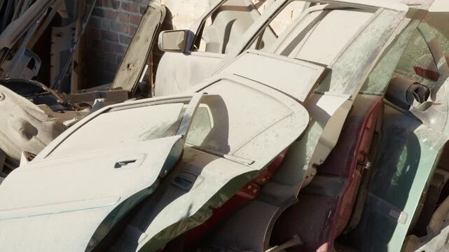 Dusty car doors stand stacked against a brick wall at an auto salvage yard. Stripped vehicle body panels await reuse or secondary metal recycling disposal.