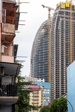 Weathered residential buildings with dangerous balconies against massive concrete skyscraper construction site with tower cranes in Batumi, Georgia, chaotic urban development, poor quality housing.