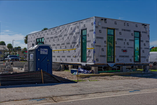 A modular home wrapped in weather barrier sits elevated on blocks at a sunny urban construction site Editorial Use Only October 16, 2026 St. Pete Beach, FL USA with a blue portable toilet, concrete bl