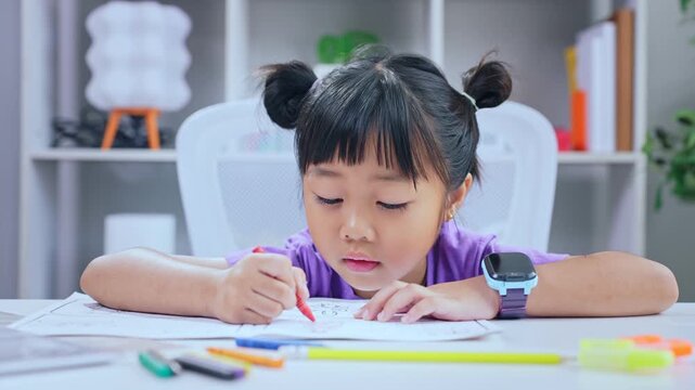 Asian girl student writing homework attentively with red pen at desk in living room for primary education concept