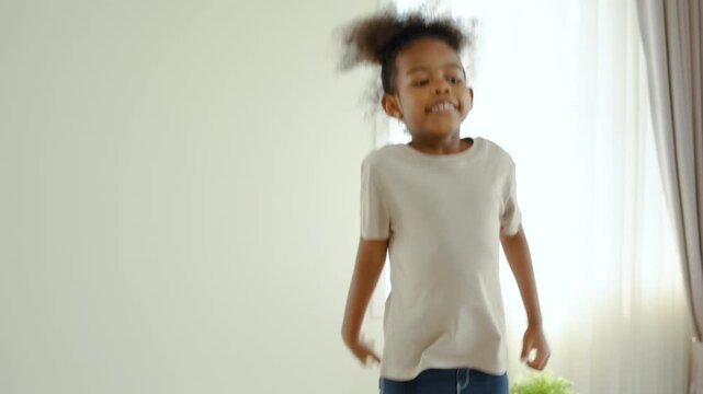 Joyful little African American girl jumping and smiling happily in a bright living room at home. Energetic cute child having fun indoors, childhood happiness, joyful lifestyle concept stock footage.