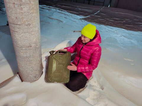 Woman holding a gasoline can while kneeling in snow beside a porch pillar. Winter fuel storage and cold weather preparedness. Household energy and emergency supply concept