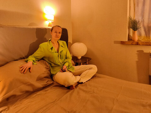 Woman sitting on bed holding television remote control in bedroom. Home entertainment and television viewing. Relaxation, leisure and evening routine. Cozy domestic lifestyle