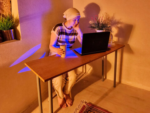 A woman wearing a headscarf works on a laptop at her desk at home. Online work and distance learning. Digital technology, internet access, and a home office. Warm, colorful interior portrait