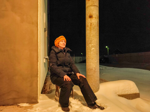 Woman sitting outdoors on a snowy porch at night. Winter weather and cold season. Snowfall, frost and freezing conditions. Outdoor winter life and seasonal survival