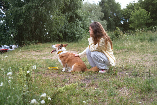 Woman sits on grass beside corgi in a park during a sunny day