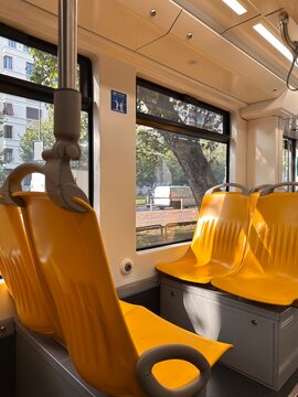 Empty yellow seats inside city tram with metal poles, hanging grips and wide windows overlooking a leafy urban street during daytime, public transport