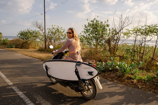 Woman riding scooter with surfboard along coastal road