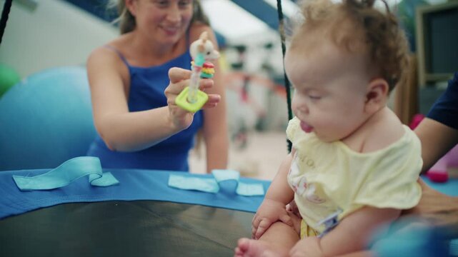 Baby girl sitting on a round therapy swing as a smiling woman shows her a colorful teether toy in a therapy room, French Riviera, France.