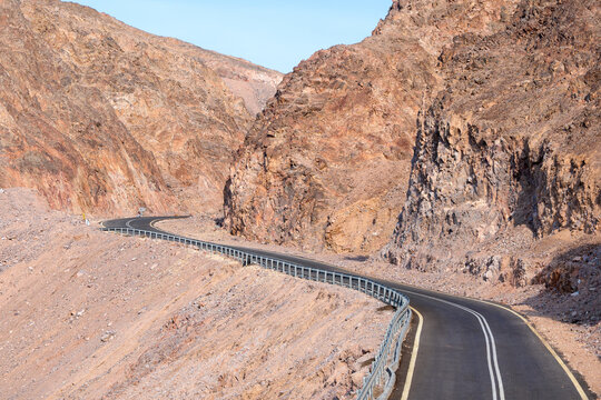 Winding asphalt road with sharp switchbacks and guardrails crosses a steep barren mountainside in Eilat Israel.