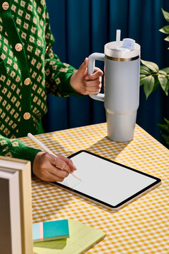 Woman's hands using stylus on tablet while holding large tumbler