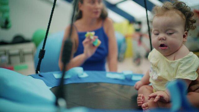 Baby girl sitting on a round platform swing looking at a teether toy held by a woman in a blue dress, French Riviera, France.