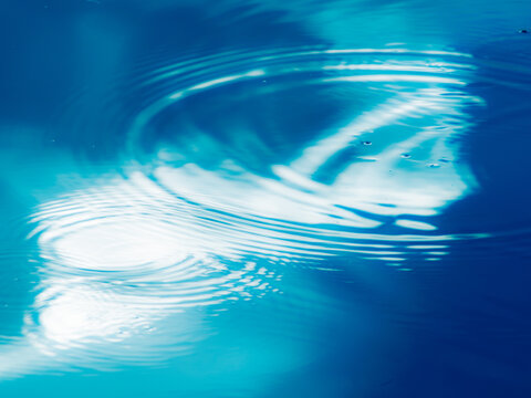 Concentric ripples spreading across vivid blue water with bright white cloud reflections, dynamic abstract water surface background