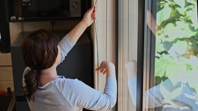 Dark haired woman pulling down roller shade in bright modern kitchen, managing heat and sunlight, summer home comfort, indoor temperature regulation, passive cooling, energy saving solutions