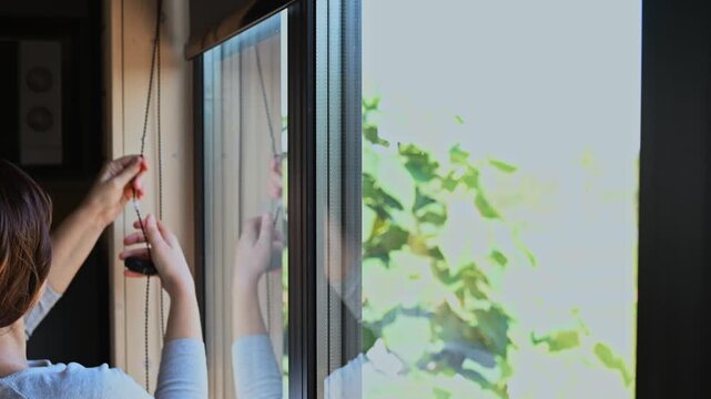 Dark haired woman operating roller blind at large kitchen window, morning or evening household routine, independent living. The problem of curious neighbors peeking into your windows