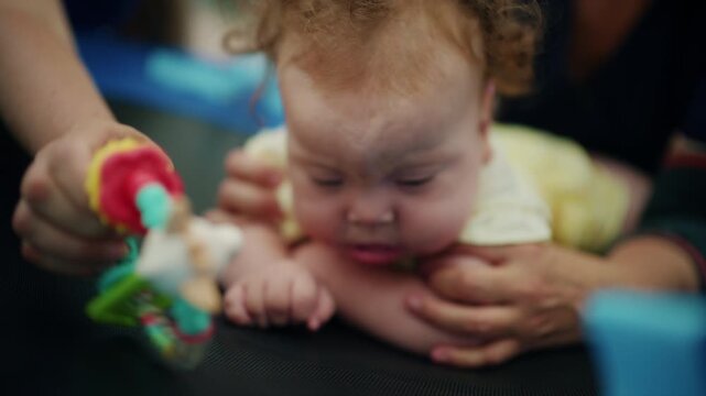 Close-up of a baby girl doing tummy time on a round therapy swing while therapist hands hold her arms and a teether toy, French Riviera, France.