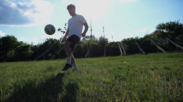 Young sportsman juggling a soccer ball on a green grass field. Male football player practicing impressive tricks and freestyle skills on stadium during a bright sunny summer day. Slow motion