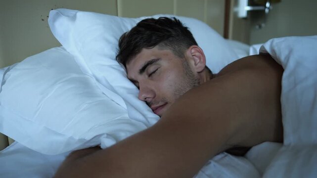 Young man suddenly waking up in bed with surprised expression in modern hotel room or home interior. Emotional guy reacting to abrupt awakening showing sleep interruption and natural human reaction