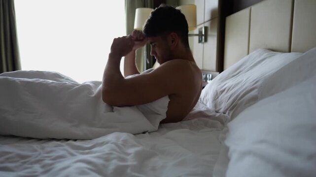 Sleepy young man waking up and sitting on edge of bed in modern hotel room or home interior after waking up. Relaxed guy enjoying quiet morning atmosphere, showing natural awakening and comfort