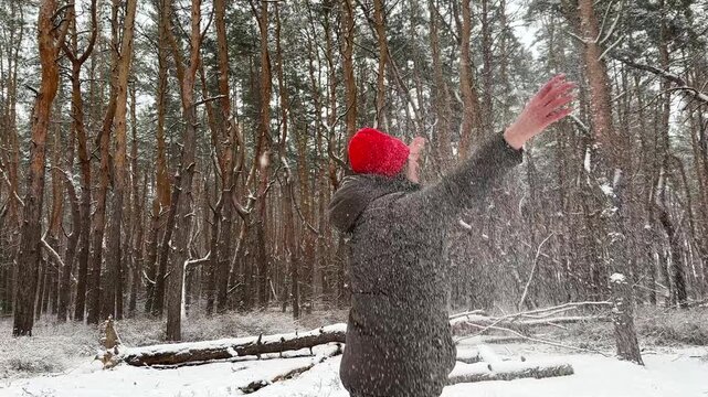 Happy adult woman throw up fresh snow and spinning under falling cloud of snowflakes at wood. Cheerful lady having fun in winter forest with joyful expression. Carefree female enjoy winter vacation