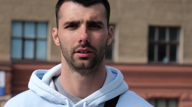 Portrait of young confident bearded man in casual clothing with cityscape background. Close up of handsome guy looks into camera standing at city environment at sunny day