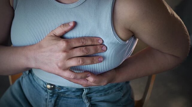 Unrecognizable woman performing a breast self-examination while sitting in an armchair at home. Breast pain, breast cancer, mastopathy, early detection of a breast tumor. Close-up of hands.