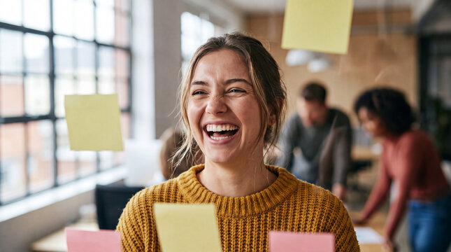 A cheerful young woman laughing during a productive creative brainstorming session in a modern office with colorful sticky notes on a glass wall.