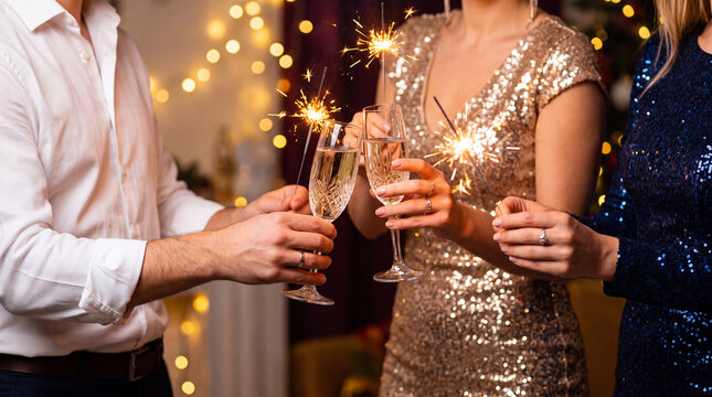 A group of friends in festive outfits holding champagne glasses and glowing sparklers during a New Year's Eve celebration with bokeh lights.