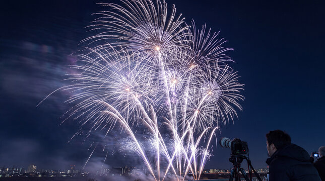 A photographer using a tripod-mounted camera to capture a brilliant display of white fireworks illuminating the night sky above a cityscape.