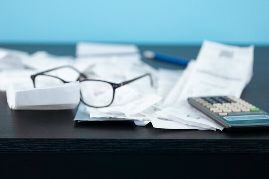 pile of shopping receipts and utility bills with glasses, pen and calculator on dark desk, blue wall with copy space for banner design, concept of household budget, finance, cost of living
