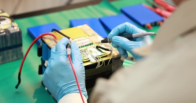 Close-up of a technician wearing blue gloves testing a battery module with probes on an electronics workbench.