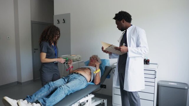 A medical team prepares a senior man for an ECG examination in a clinic. A female healthcare worker attaches wrist electrodes while a male doctor reviews a medical folder.