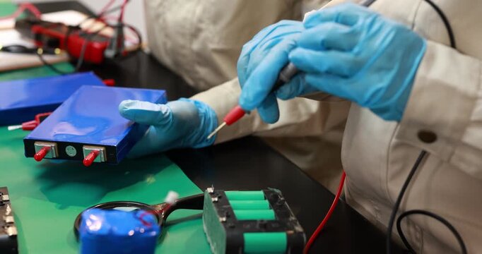 Technician in blue gloves testing an electronic battery module with probes at a workbench in a clean manufacturing lab.