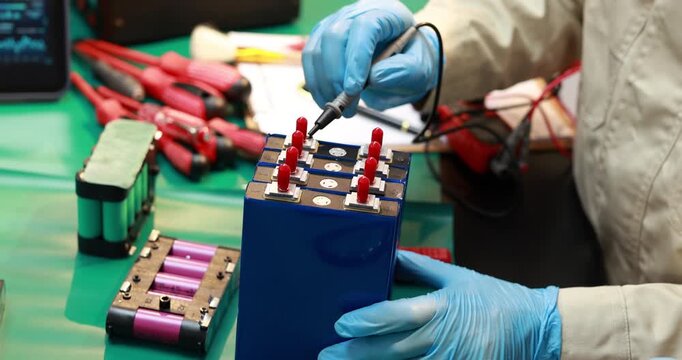 Technician in protective gloves tests a lithium battery pack with probes at an electronics workbench, surrounded by tools and battery cells