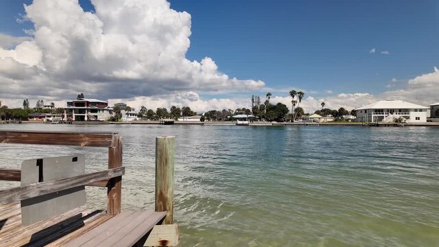 A wooden boardwalk runs beside a calm coastal canal in Treasure Island,FL as dramatic storm clouds gather overhead, with waterfront homes, docks and a small boat visible in the distance, conveying ten