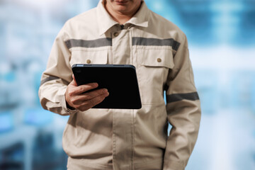 Technician in beige work uniform holding a tablet in a blurred industrial workplace, showing digital inspection, maintenance, and modern field service. © Phushutter