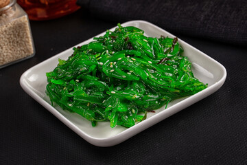 White square ceramic plate holding bright green chuka seaweed salad with sesame seeds on dark table surface © Andrei Starostin
