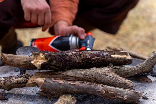 A person uses a portable gas torch to ignite stacked firewood, directing the burner nozzle at branches arranged for an outdoor campfire.