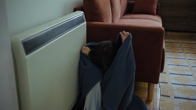 Freezing girl wrapped in a warm knitted blanket sitting on the floor between the sofa and electric radiator, trying to warm up in a cold apartment during winter.