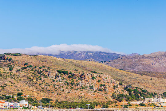 Beautiful view of rocky mountains with low clouds above coastal town and harbor. Crete. Greece.